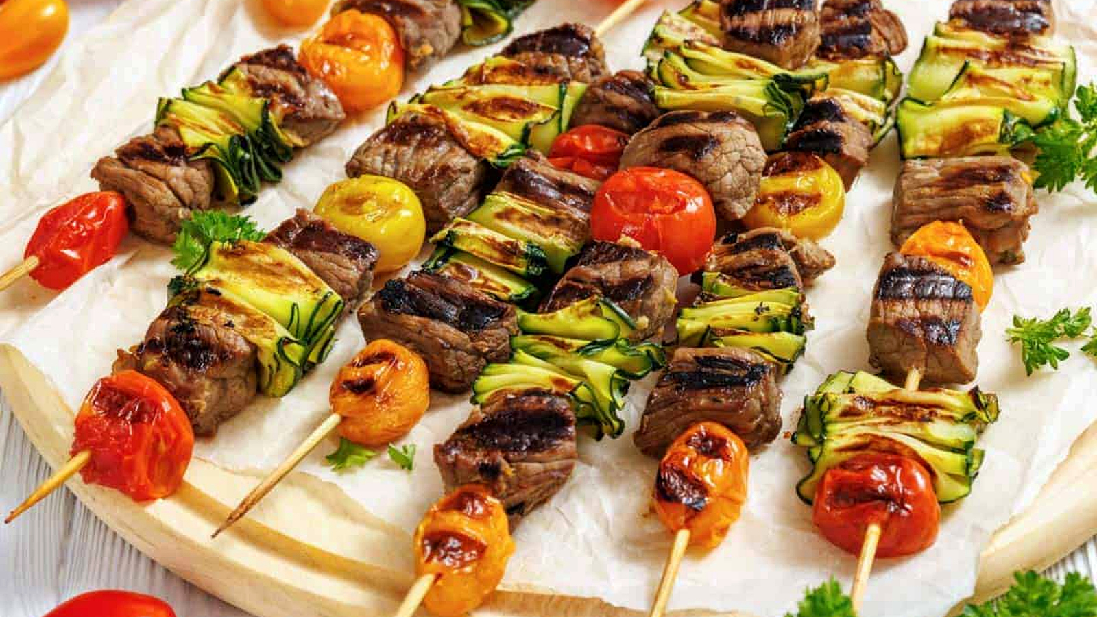 A wooden round serving board is covered with white parchment paper, displaying numerous grilled steak and veggie skewers arranged horizontally across it.