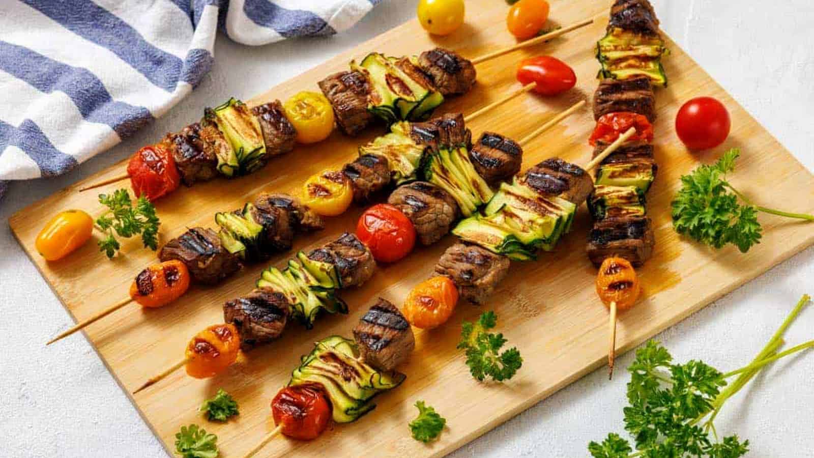 Grilled skewers with beef, zucchini, and cherry tomatoes on a wooden board, garnished with fresh parsley. A striped cloth and loose cherry tomatoes are nearby.