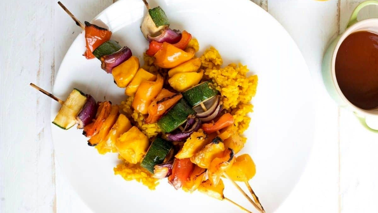 Three Grilled Teriyaki Mango Skewers with bell peppers, zucchini, and onions served on a bed of yellow rice on a white plate.