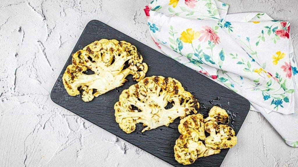 Three grilled cauliflower steaks on a black rectangular plate, next to a white napkin with a colorful floral pattern.
