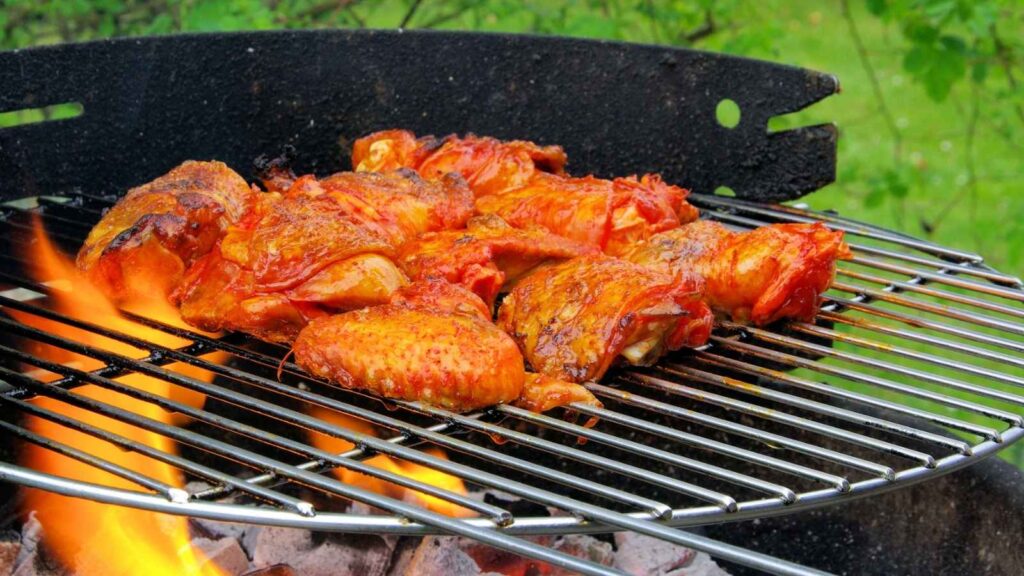 Chicken pieces being grilled over an open flame on a barbecue, with some pieces already browned and charred&mdash;perfect for putting your favorite grilling tips for foods to use&mdash;outdoors in a lush, green setting.