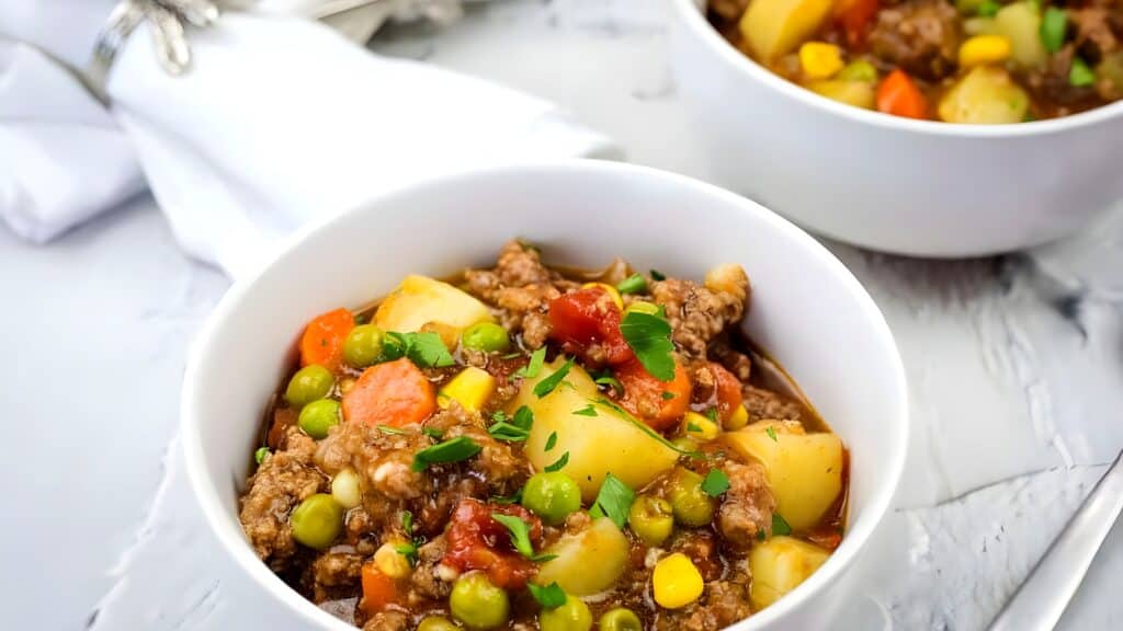 A white bowl filled with beef stew containing potatoes, peas, carrots, corn, and a tomato-based broth, garnished with chopped herbs.