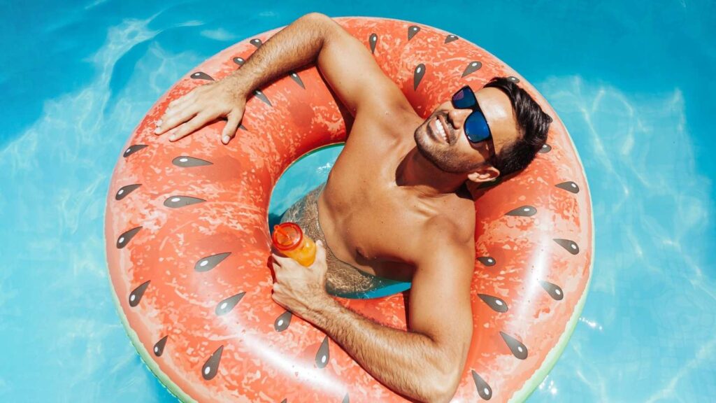 A man wearing sunglasses relaxes in a pool on a watermelon-shaped inflatable ring, holding a drink bottle.