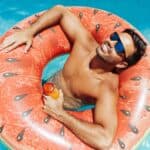 A man wearing sunglasses relaxes in a pool on a watermelon-shaped inflatable ring, holding a drink bottle.
