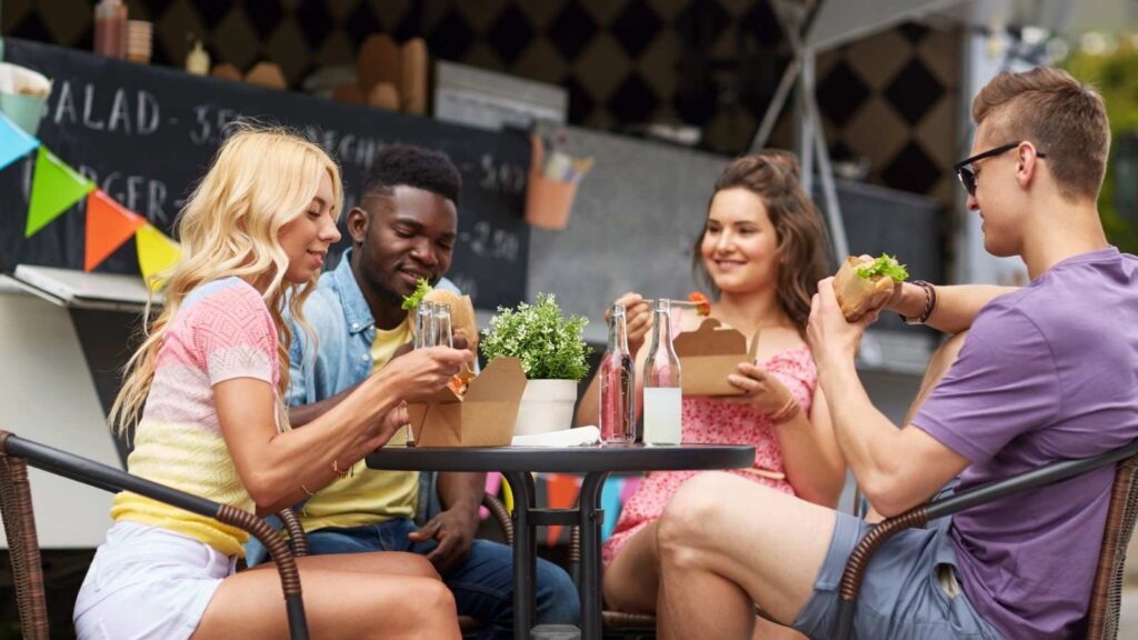 Four people sit at an outdoor table enjoying takeout, drinks, and a small potted plant centerpiece. A food truck in the background sets the scene for a food-forward travel experience.