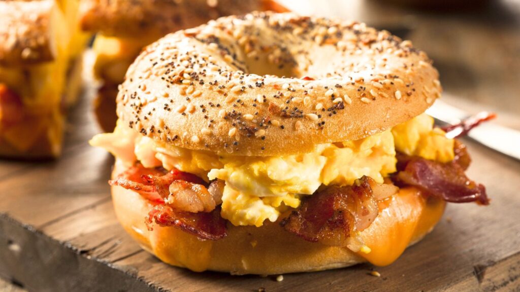 An everything bagel sandwich filled with scrambled eggs, crispy bacon, and melted cheese sits on a wooden surface&mdash;a tempting choice for anyone with an expensive breakfast habit.