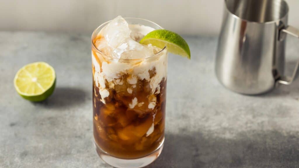 A glass of iced coffee with cream is topped with ice and garnished with a lime wedge. A halved lime and a metal pitcher are in the background.