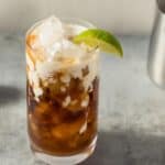 A glass of iced coffee with cream is topped with ice and garnished with a lime wedge. A halved lime and a metal pitcher are in the background.