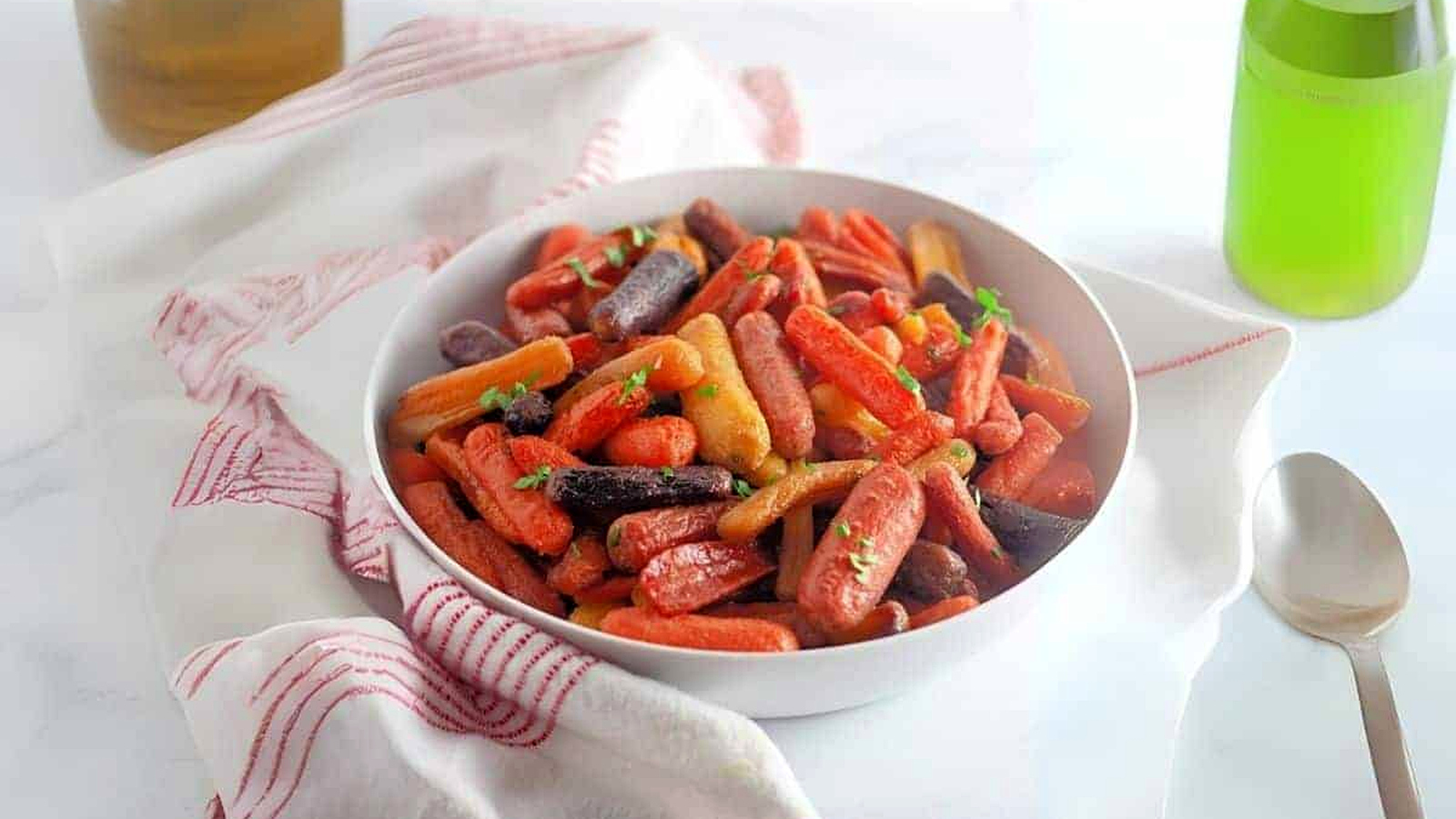 A white bowl filled with honey-roasted baby carrots sits on a white cloth with red stripes, next to a spoon and a green bottle on a light surface.