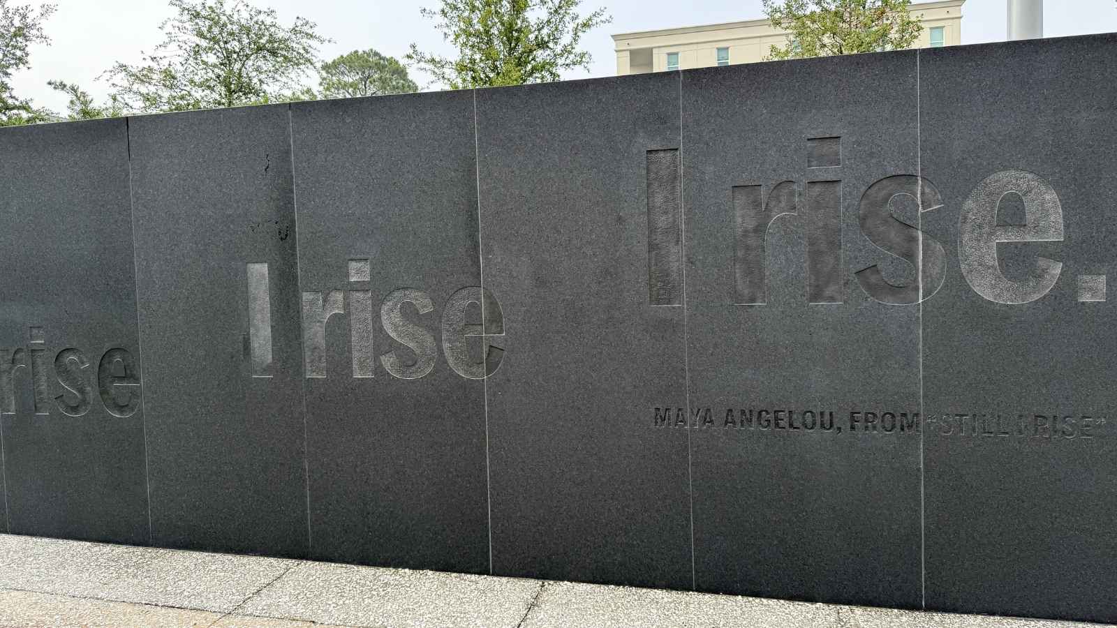 Black stone wall engraved with the words “I rise. I rise.” and “Maya Angelou, from ‘Still I Rise,’” with trees and a building visible in the background.