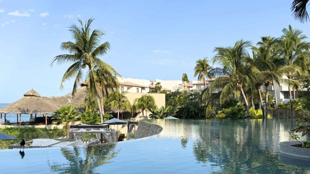 Infinity pool with clear water in the foreground, surrounded by palm trees, resort buildings, thatched huts, and blue sky in the background.