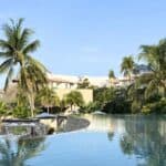 Infinity pool with clear water in the foreground, surrounded by palm trees, resort buildings, thatched huts, and blue sky in the background.