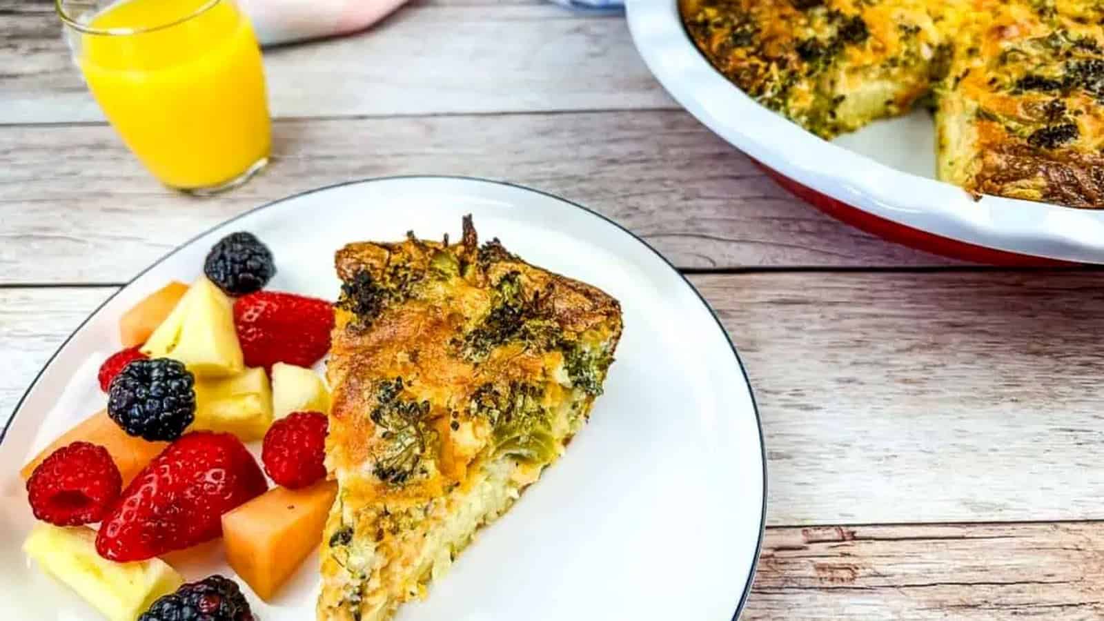 A slice of Broccoli 3-Cheese Impossible Quiche on a white plate with a side of mixed fruit. A full quiche and glass of orange juice are in the background.