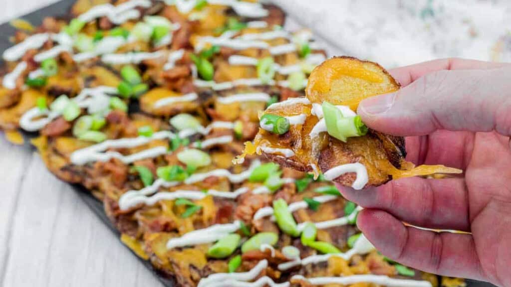 A hand holds a loaded potato chip topped with melted cheese, sour cream, and chopped green onions; a tray of similar chips is in the background.
