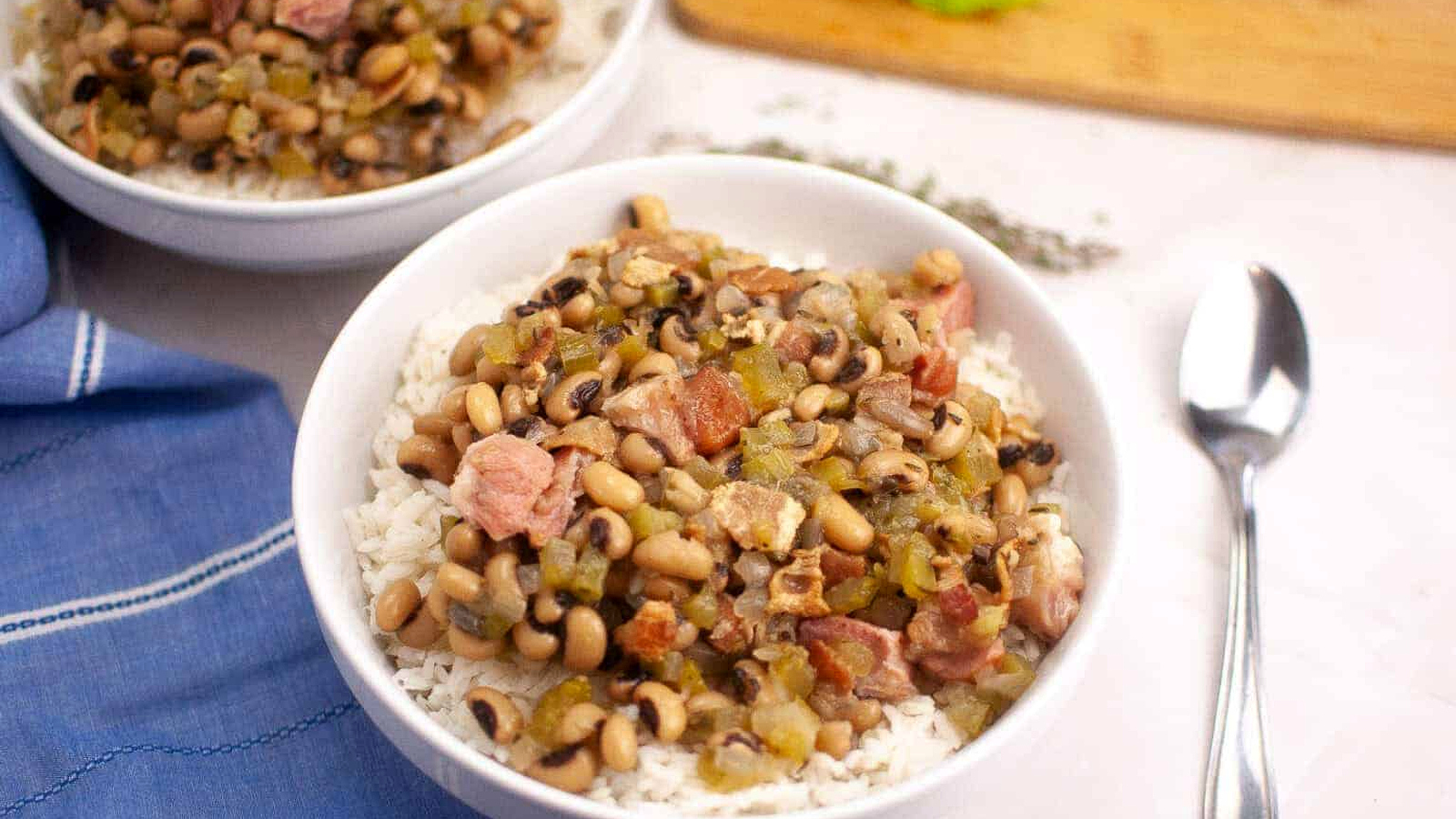 A bowl of Hoppin&rsquo; John served over white rice, with a spoon and a blue napkin nearby.