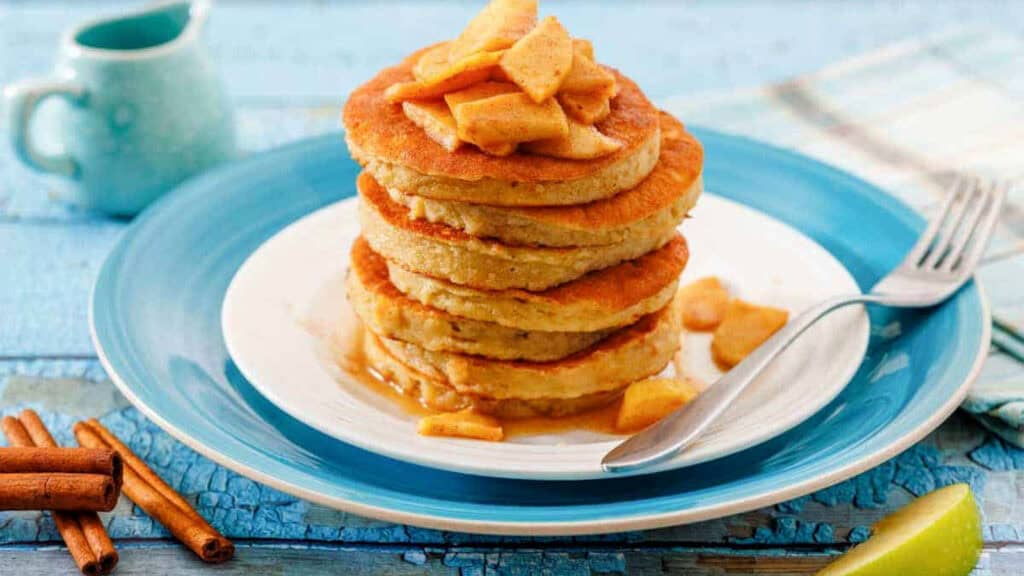 A stack of pancakes topped with cooked apple slices and syrup is served on a white plate with a fork, set on a blue table. Cinnamon sticks and a napkin are nearby.