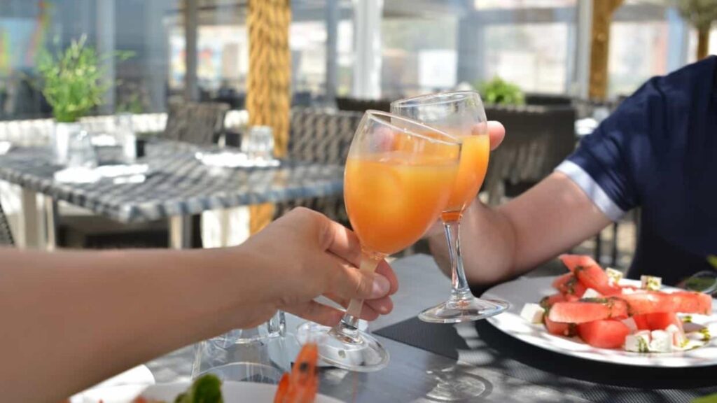 Two people enjoying daytime drinking, clinking glasses of orange juice at an outdoor dining table with plates of food, including watermelon salad, in the background.