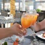 Two people enjoying daytime drinking, clinking glasses of orange juice at an outdoor dining table with plates of food, including watermelon salad, in the background.