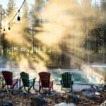 Four Adirondack chairs face a steaming outdoor pool surrounded by trees, with sunlight streaming through mist in a forested setting.