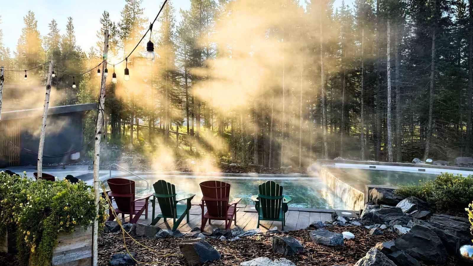 Four Adirondack chairs face a steaming outdoor pool surrounded by trees, with sunlight streaming through mist in a forested setting.