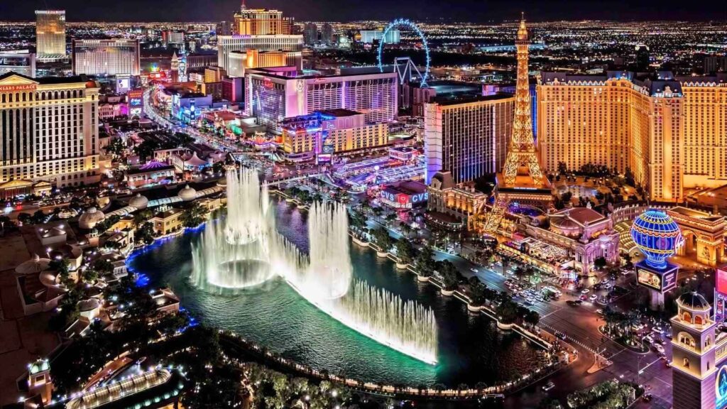 Aerial view of the Las Vegas Strip at night with illuminated hotels, casinos, the Eiffel Tower replica, and the Bellagio fountains in action.