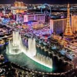 Aerial view of the Las Vegas Strip at night with illuminated hotels, casinos, the Eiffel Tower replica, and the Bellagio fountains in action.