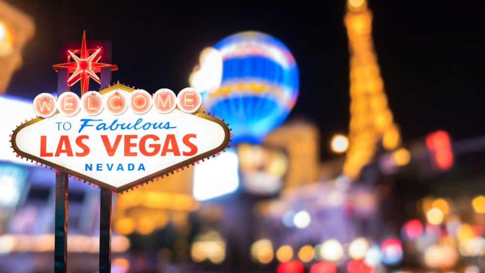 The illuminated Welcome to Fabulous Las Vegas, Nevada sign stands in front of blurred colorful lights and landmarks at night.