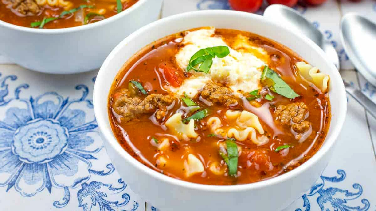 Close-up of a bowl of soup with lasagna soup, topped with chopped basil, on a blue-patterned tiles.