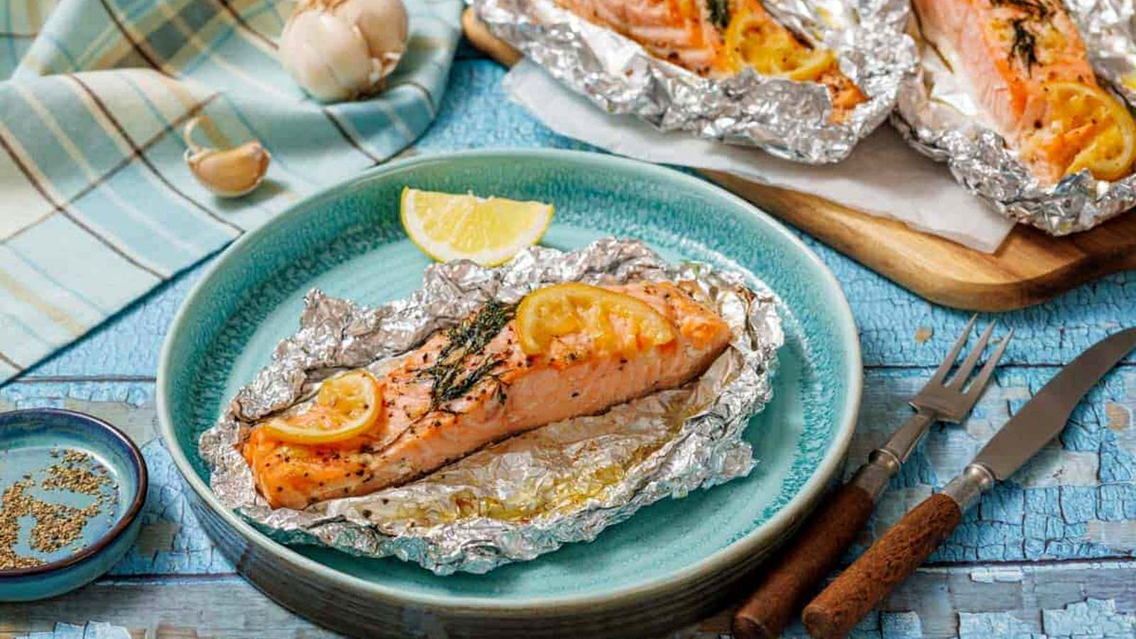 Lemon-Dill Salmon Foil Packets and herbs served in aluminum foil on a blue plate, with utensils and garlic nearby.