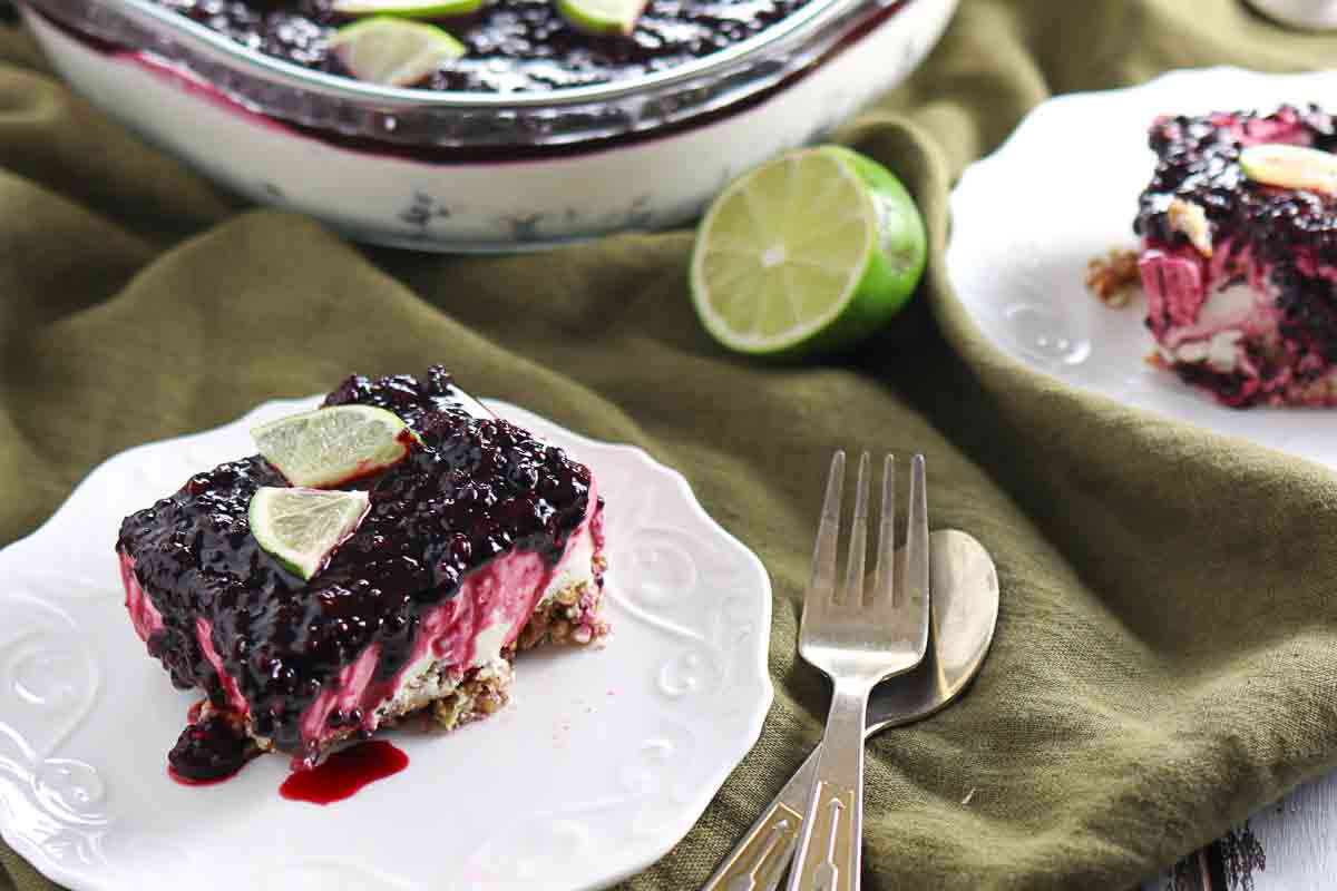 A slice of berry-topped dessert with lime wedges sits on a white plate next to a fork and knife, with more dessert and a halved lime in the background.