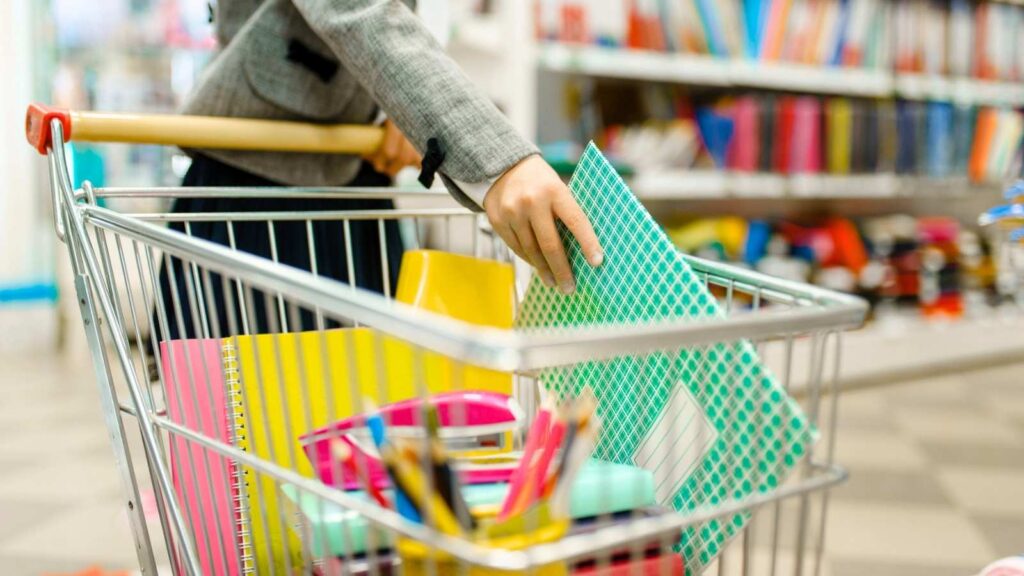 A person places notebooks into a shopping cart filled with school supplies in a store aisle.