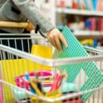 A person places notebooks into a shopping cart filled with school supplies in a store aisle.