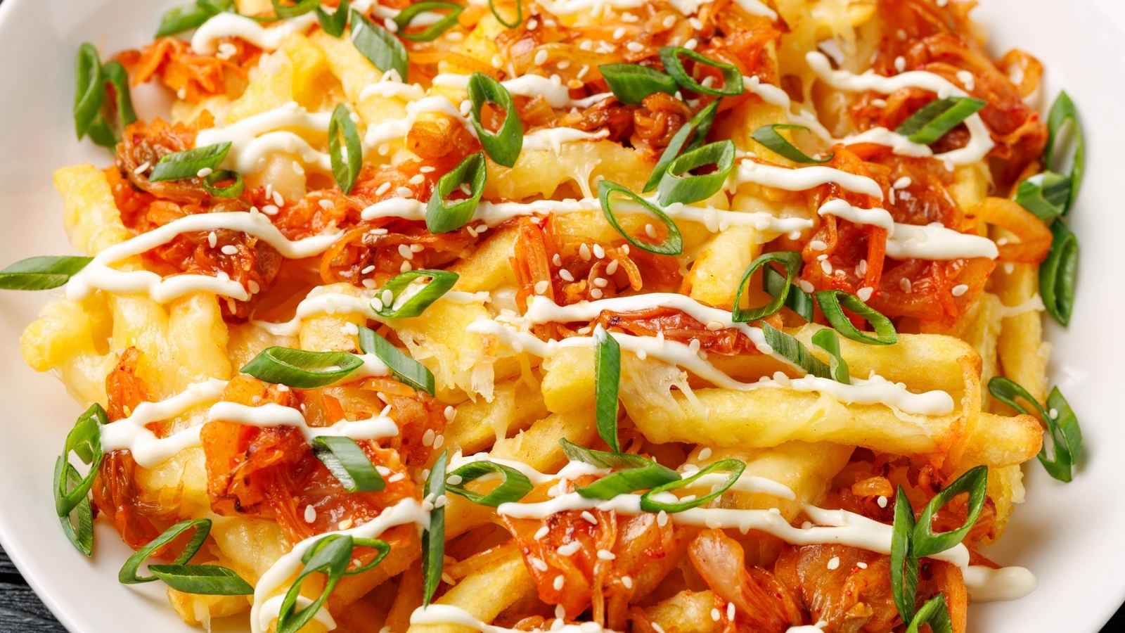 A plate of French fries topped with kimchi, sliced green onions, sesame seeds, and drizzled with mayonnaise.