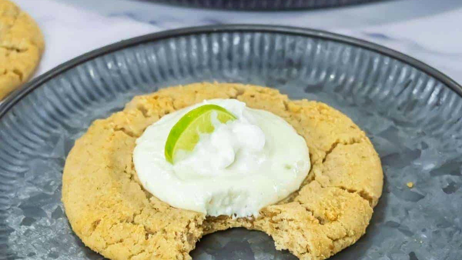 A large Copycat Crumbl Key Lime Cookies with a bite taken out, topped with white cream and a lime wedge, served on a gray plate.