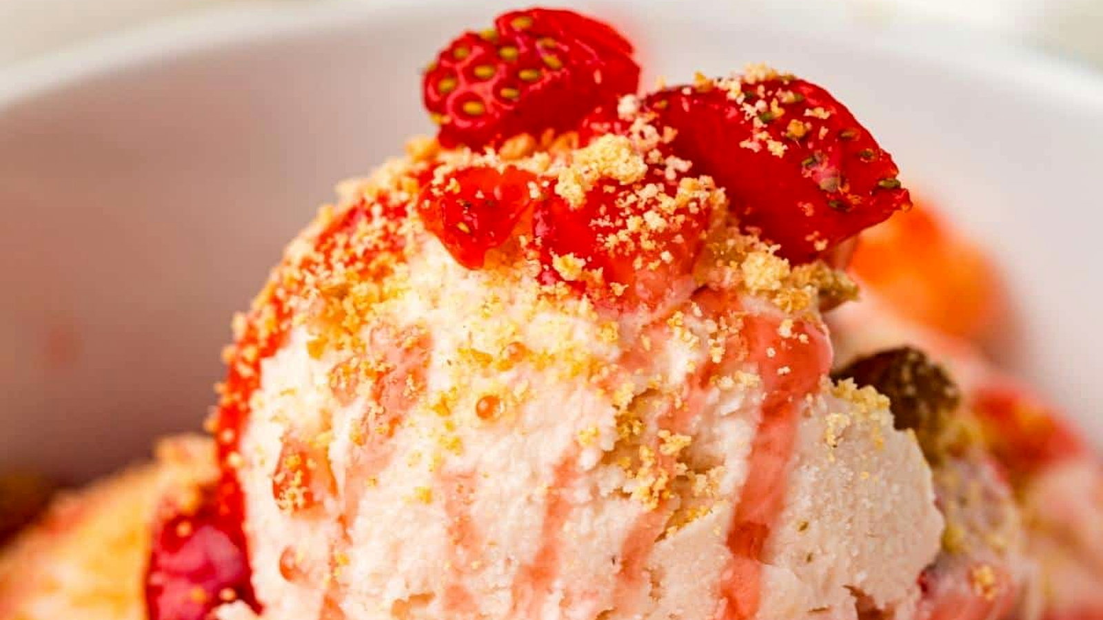Close-up of a bowl of ice cream topped with crushed cookies, strawberry pieces, and a drizzle of strawberry sauce.