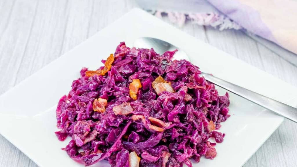 A serving of braised red cabbage with pieces of bacon on a square white plate, accompanied by a metal spoon.