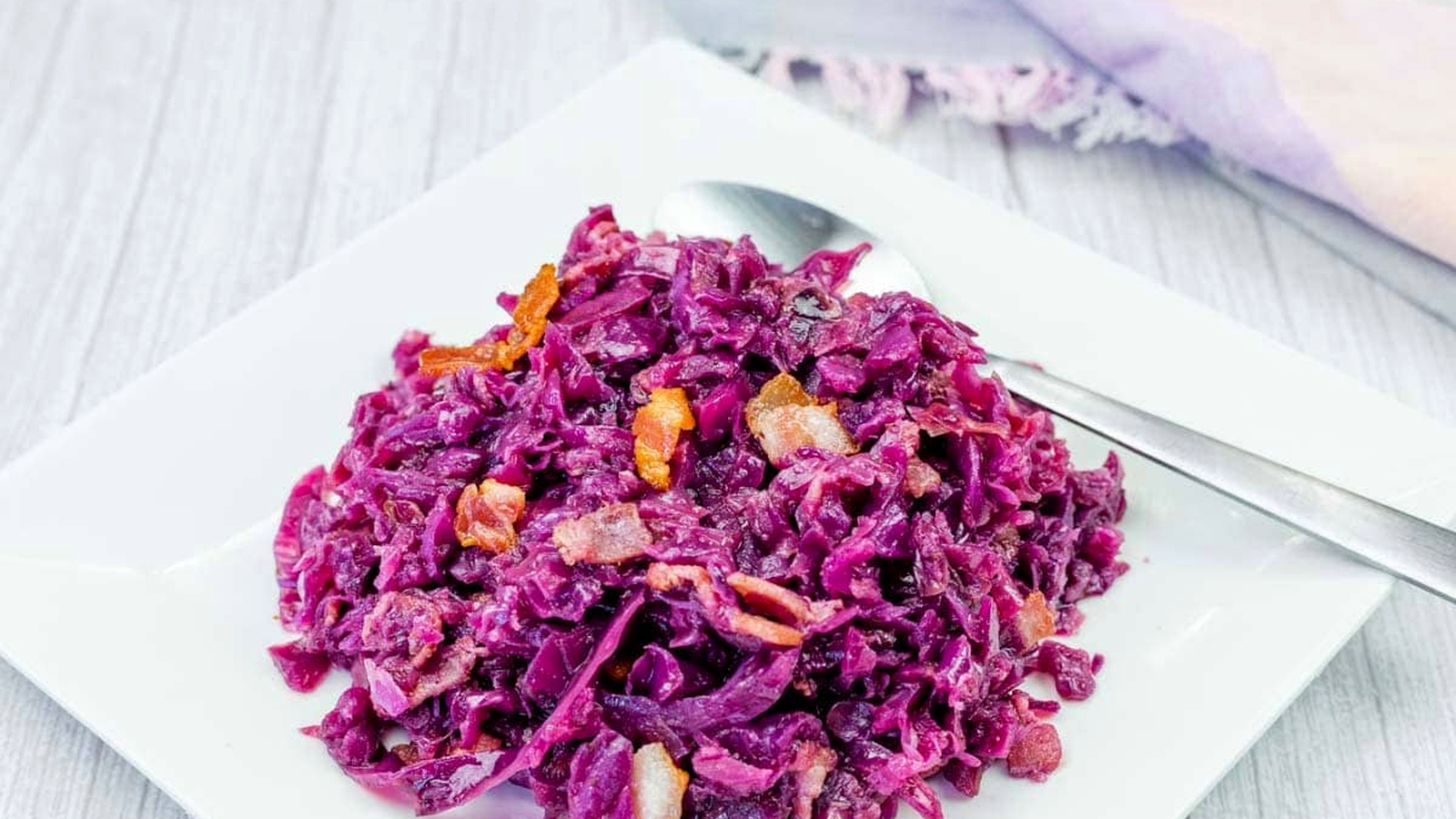 A serving of cooked red cabbage with pieces of bacon and carrots on a white square plate, with a spoon beside the food.