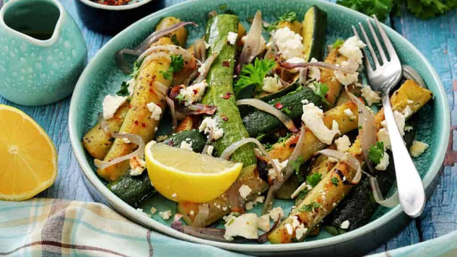 A plate of grilled zucchini with crumbled feta, sliced onions, fresh parsley, and a lemon wedge, served on a blue table with a fork and a checkered napkin.