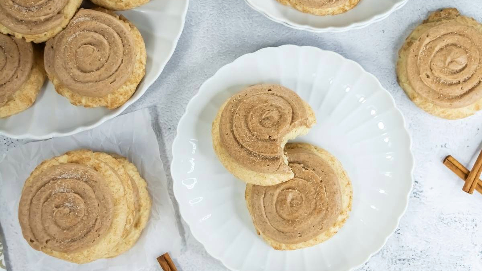 Crumbl Copycat Churro Cookies are arranged on white plates and a napkin. One cookie on a plate has a bite taken out. Two cinnamon sticks are placed nearby on the white surface.