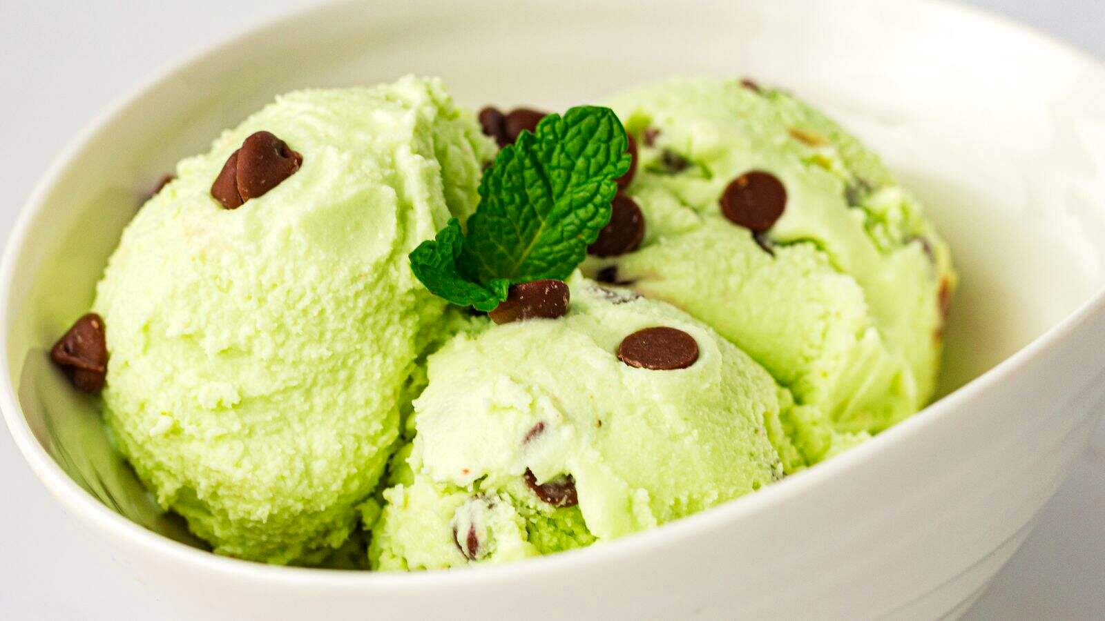 Three scoops of mint chocolate chip ice cream in a glass bowl, with chocolate pieces visible and a sprig of mint in the background.
