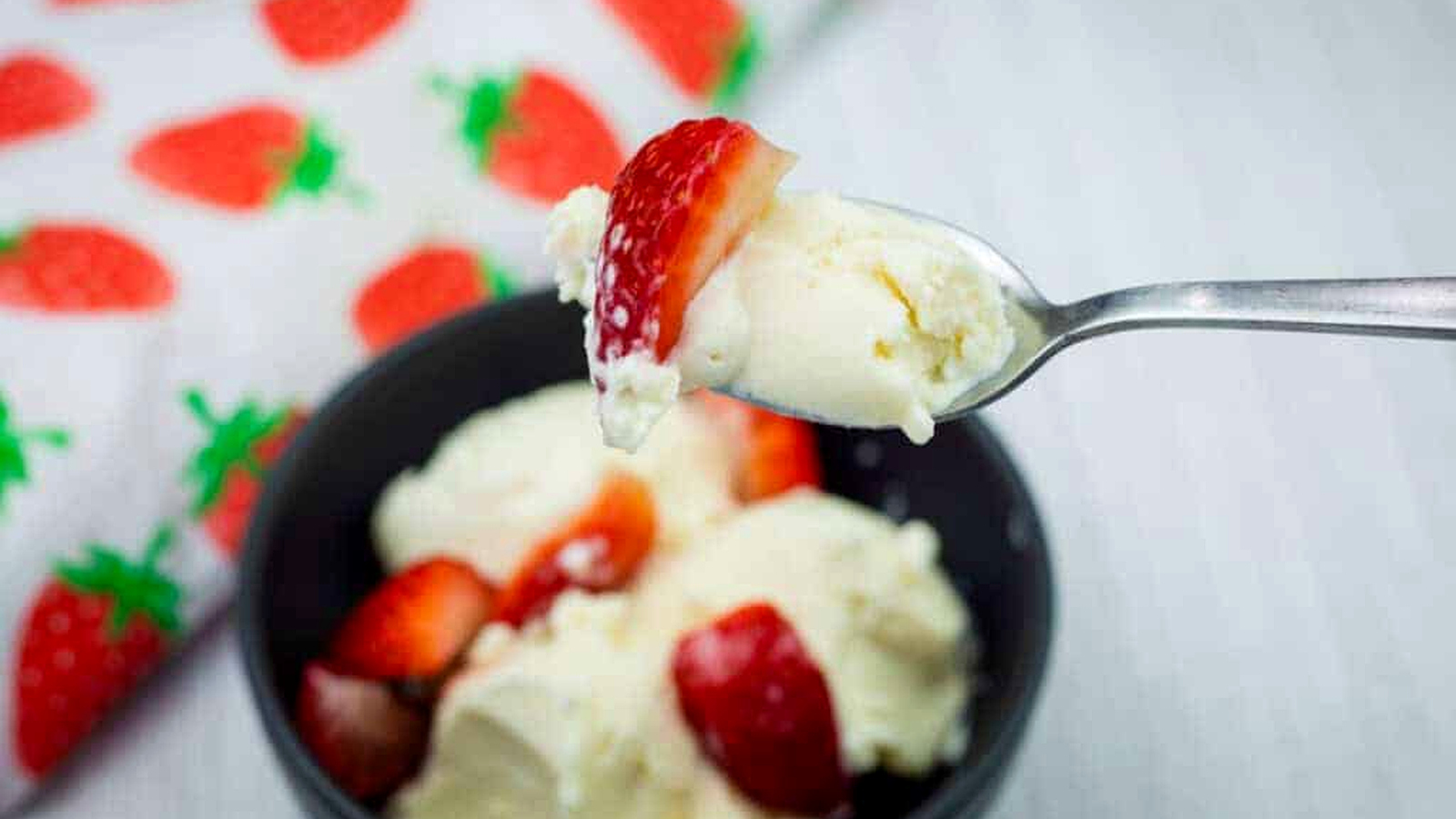 A spoonful of vanilla ice cream and a strawberry held above a bowl with more ice cream and strawberries, next to a strawberry-patterned cloth.