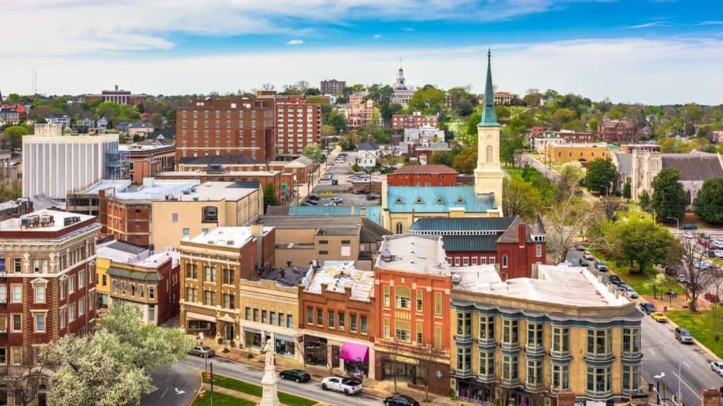 Aerial view of Macon, Georgia&rsquo;s downtown with historic buildings, a church steeple, tree-lined streets, and a courthouse dome visible in the distance beneath a partly cloudy sky.