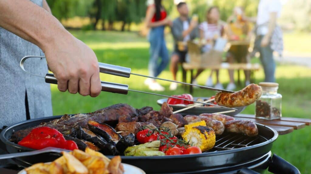 Person grilling various meats and vegetables on a barbecue outdoors with a group of people sitting at a picnic table in the blurred background.