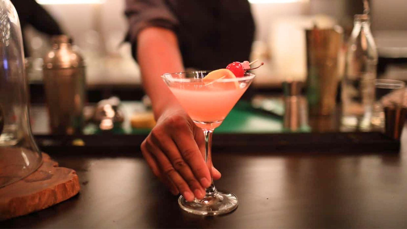 A bartender places a pink cocktail garnished with a lemon slice and cherry on a bar counter.