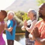 Four adults stand outdoors in a row, eyes closed and hands in prayer position, peacefully practicing meditation or yoga on a sunny International Day of Yoga.