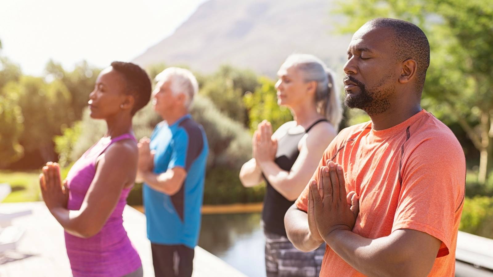 Four adults stand outdoors in a row, eyes closed and hands in prayer position, peacefully practicing meditation or yoga on a sunny International Day of Yoga.