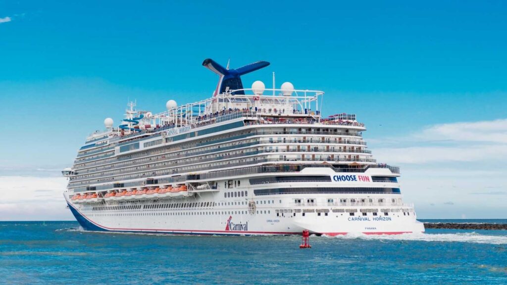 A large white Carnival cruise ship sails on blue water near the shore under a clear sky, with &ldquo;CHOOSE FUN&rdquo; visible on the side.