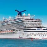 A large white Carnival cruise ship sails on blue water near the shore under a clear sky, with “CHOOSE FUN” visible on the side.