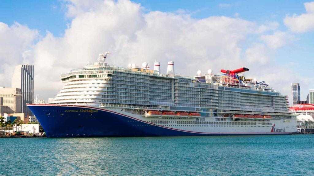 A large white cruise ship, the Carnival Celebration, is docked at a port with city buildings and a partly cloudy sky in the background.