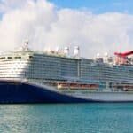 A large white cruise ship, the Carnival Celebration, is docked at a port with city buildings and a partly cloudy sky in the background.